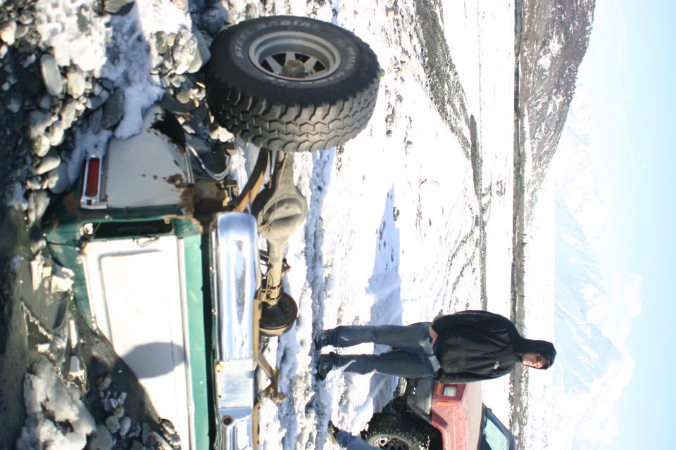 Person standing next to a green off-road vehicle stuck in snow and mud, with another red vehicle partially visible