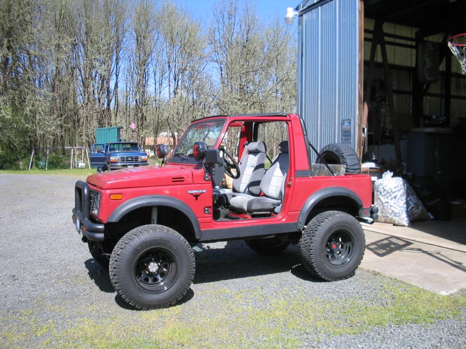 Red Suzuki Samurai with aftermarket wheels and tires, spare tire mounted at rear, open doors, parked on gravel