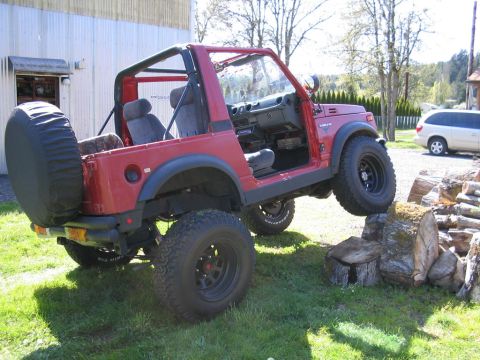 1987 Suzuki Samurai - A red Suzuki Samurai is shown off-road, with one tire on a log, next to a white minivan.