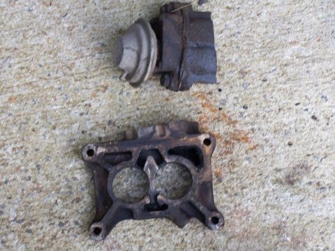 1979 Ford F-150 - Two engine parts, a carburetor base and a vacuum actuator, lie on a textured concrete surface.