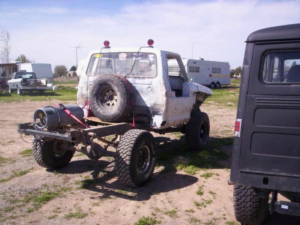 1978 Ford Bronco chassis with spare tire mounted on back and a gas tank strapped to the frame. Another vehicle is parked to the right