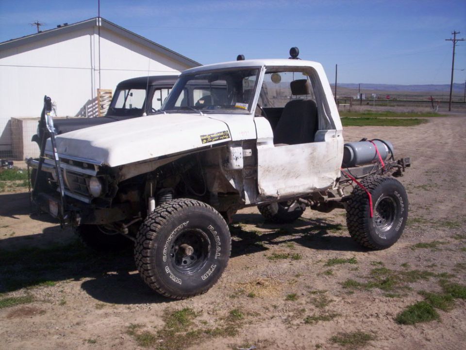 White pickup truck with no bed, large tires, and a gas tank mounted on the back. Another dark pickup truck is parked behind it
