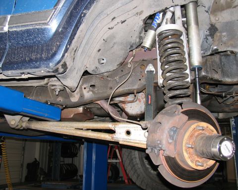 1984 Ford Bronco - Wheel-off underside view of a dark blue truck on a shop lift, showing the front coil spring, radius arm, Fox remote-reservoir shock and a n…