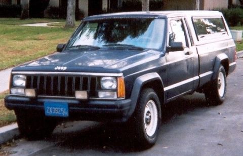 1986 Jeep Comanche - A dark blue Jeep pickup truck with a camper shell is parked curbside, showing its front and side profile.