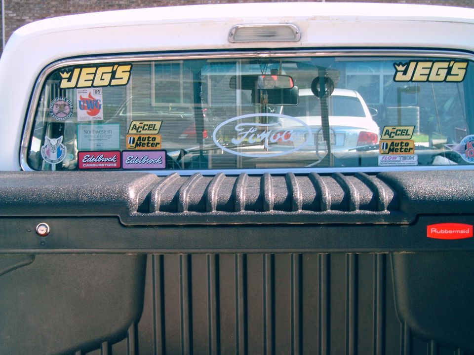 White Ford F-100 pickup truck rear window with various stickers, including JEG's and Ford oval. Another car is visible through the window