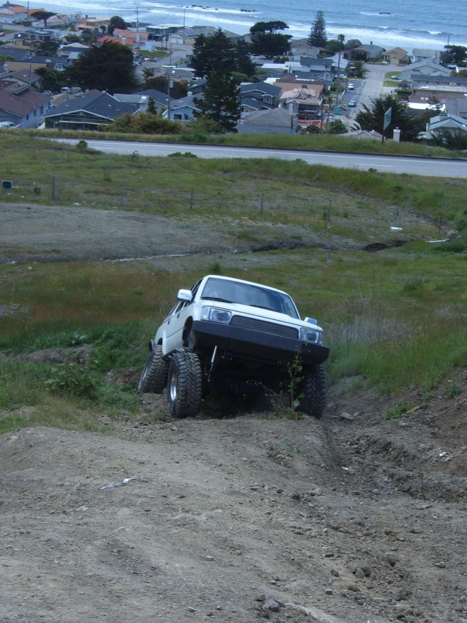 White lifted Toyota pickup on a steep dirt slope above a coastal town and the ocean