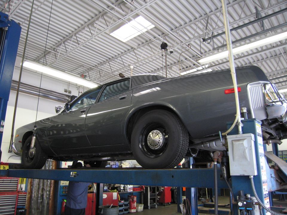 Dark gray sedan raised on a blue shop lift in a service garage, side/rear view.