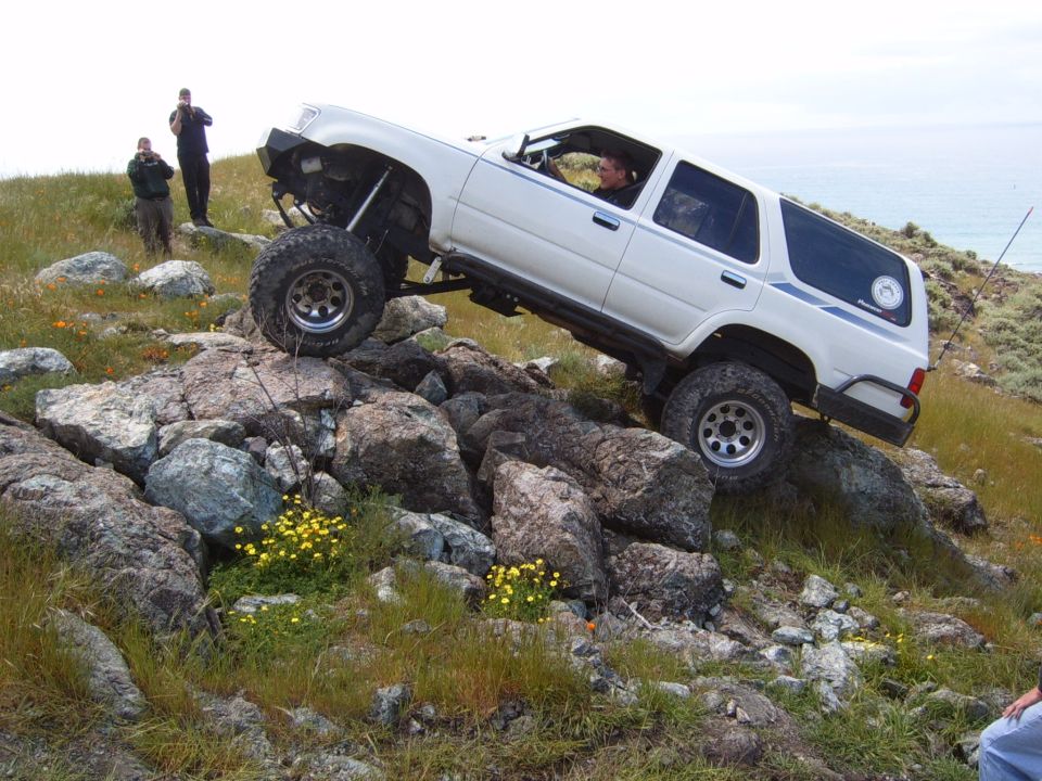 White Toyota 4Runner flexed over rocks on a grassy seaside hill with people watching