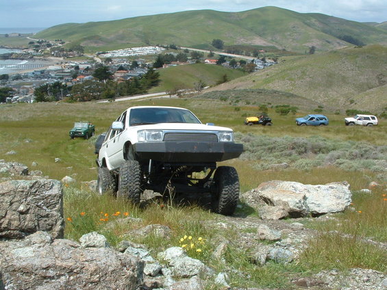 White lifted Toyota truck with custom front bumper perched on rocks above green coastal hills