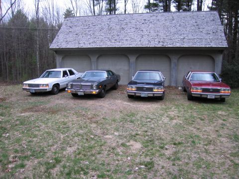 1991 Ford LTD Crown Victoria - Four sedans are parked in front of a multi-bay garage. The vehicles appear to be from the 1970s and 1980s.