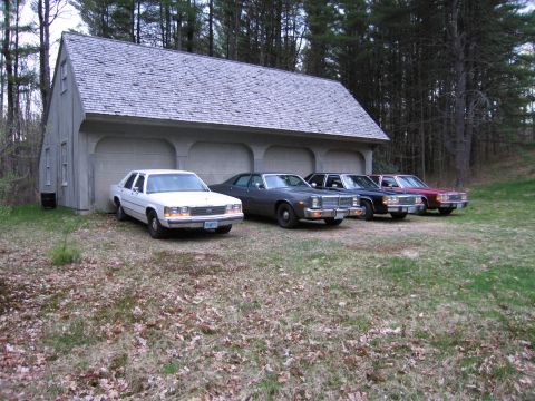 1991 Ford LTD Crown Victoria - A row of sedans, including a white Ford, are parked outside a garage.