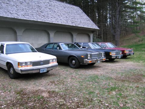 1991 Ford LTD Crown Victoria - A row of vintage sedans are parked outside a garage, showing off their distinct front-end designs.
