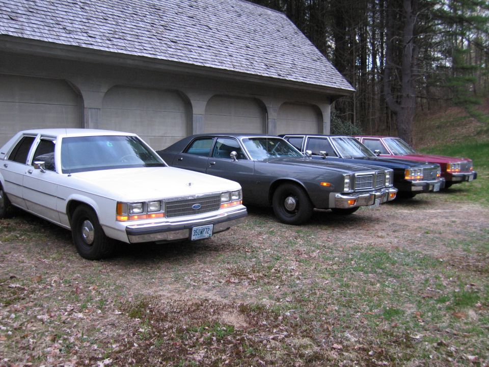 White 1980s sedan in front, followed by gray, blue, and red sedans parked in front of a garage