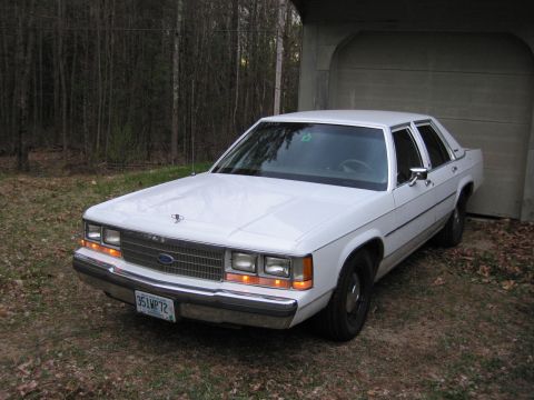 1991 Ford LTD Crown Victoria - A white sedan is parked outside a garage with trees in the background.