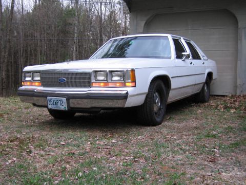 1991 Ford LTD Crown Victoria - This white sedan, likely a 1990 model, is parked in front of a garage with trees in the background.