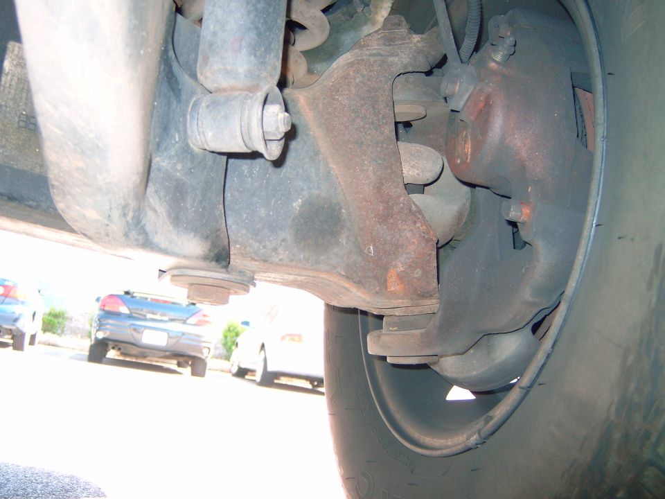 Underside view of a vehicle's front suspension and brake caliper, with other cars visible in the background