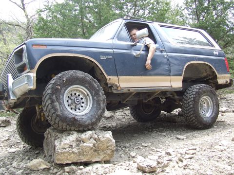 1986 Ford Bronco - A lifted blue Ford Bronco navigates rocky terrain with a driver visible.