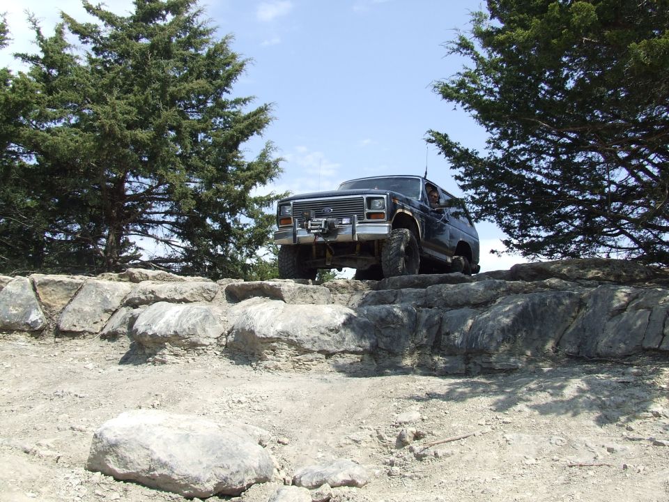 Dark blue Ford Bronco climbing over rocks on a dirt trail, trees on either side