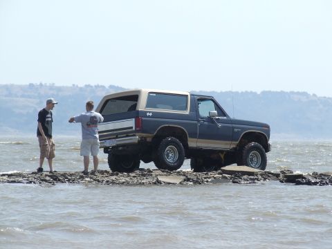 1986 Ford Bronco - A Ford Bronco sits on a rocky outcrop in the water with two people observing it.