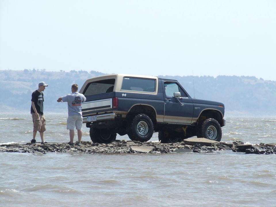 Dark blue Ford Bronco with tan top on rocky shore, two guys standing nearby