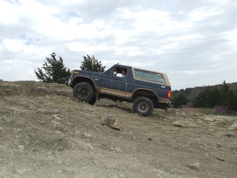 1986 Ford Bronco - A blue 1986 Ford Bronco navigates a challenging off-road course on a cloudy day.