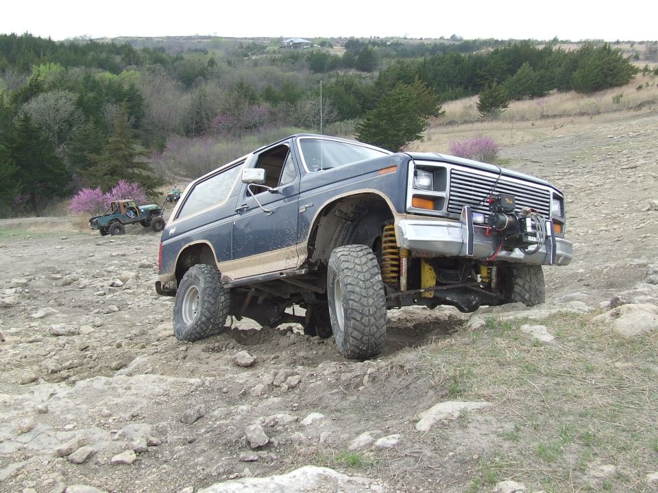 Dark blue Ford Bronco with lifted suspension and large tires driving on a rocky trail. Another green Bronco is visible in the background