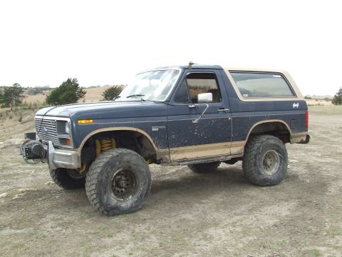 1986 Ford Bronco - A modified Ford Bronco sits on a dirt lot, its lifted suspension and winch indicating off-road use.