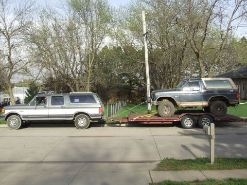 1986 Ford Bronco - A gray Ford Bronco II tows a trailer carrying a dark blue Ford Bronco, both viewed from the side.