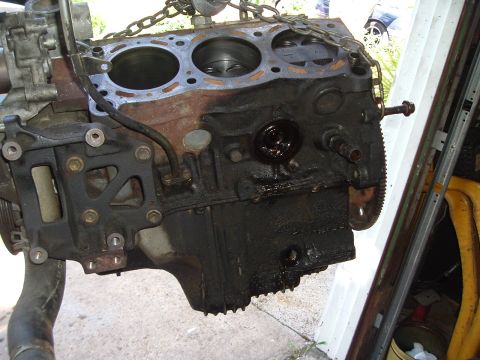 1993 Ford Taurus - An engine block, covered in oil, is suspended by chains, with a glimpse of a yellow object and shelving behind it.