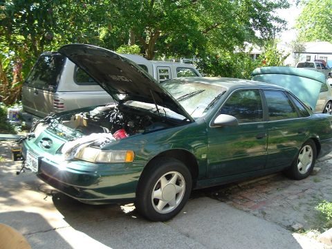 1993 Ford Taurus - Green 1993 Ford Taurus with hood open, parked in a driveway with other vehicles present.