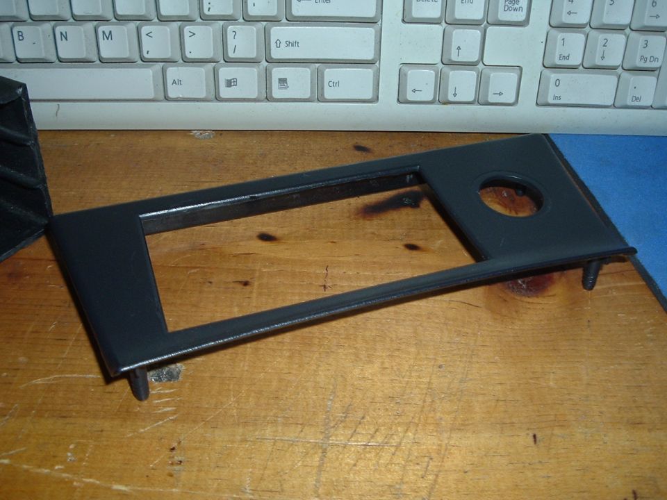 Black plastic trim piece with rectangular and circular openings, resting on a wooden surface