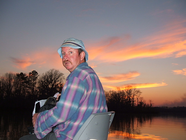 Man in a bucket hat and plaid shirt sits on a boat with a sunset in the background