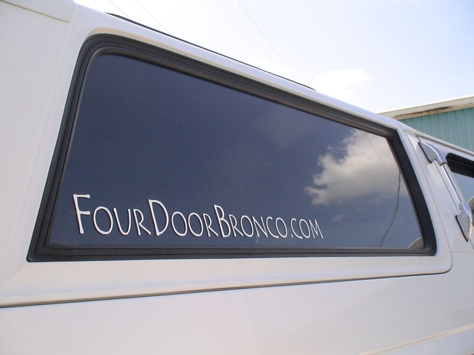 White vinyl 'FourDoorBronco.com' lettering on the tinted rear quarter window of a white vehicle