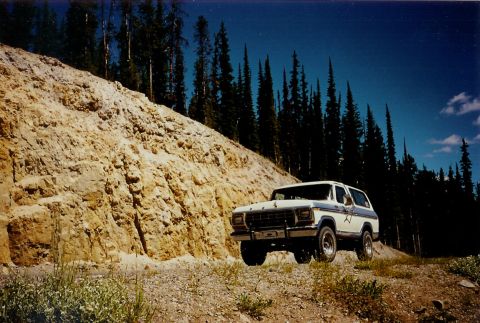 1979 Ford Bronco - A white 1978 Ford Bronco drives on a dirt road through a wooded area with a large rock face.