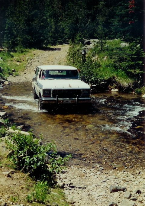 1979 Ford Bronco - White 1978 Ford Bronco navigates a shallow stream on a dirt path surrounded by trees.
