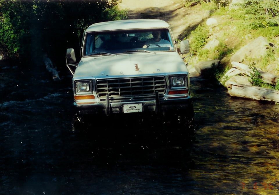 White Ford Bronco driving through a shallow stream. The grille and headlights are visible