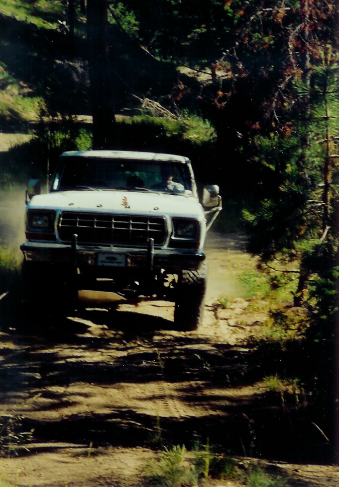 White 1979 Ford Bronco driving on a dirt road, front view. Off-road bumper and tires are visible