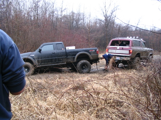 1978 Ford Bronco is stuck in mud and being pulled by a black Ford Ranger. A person is connecting the tow strap