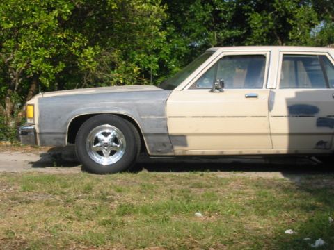1985 Ford LTD Crown Victoria - Side view of a 1985 Ford Crown Victoria LTD undergoing repairs, showing a primer fender and aftermarket wheels.