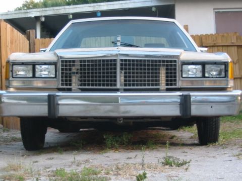 1985 Ford LTD Crown Victoria - The front of a white 1985 Ford Crown Victoria LTD is visible, its distinctive chrome grille and dual rectangular headlights.