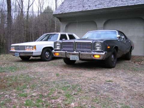 1991 Ford LTD Crown Victoria - A dark 1977 Plymouth Gran Fury is parked in front of a garage, with a white Ford sedan behind it.