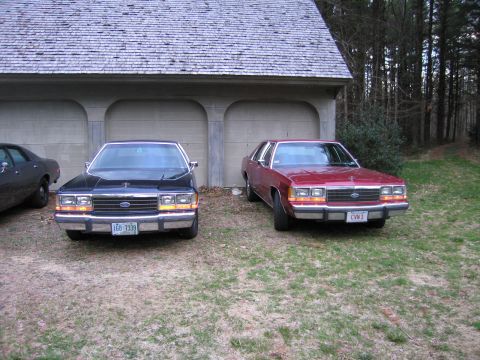 1991 Ford LTD Crown Victoria - Two Ford Crown Victorias, a black one and a red one, are parked in front of a garage.