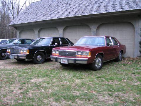 1991 Ford LTD Crown Victoria - Multiple Ford Crown Victorias are parked in front of a garage, with the closest one being maroon.
