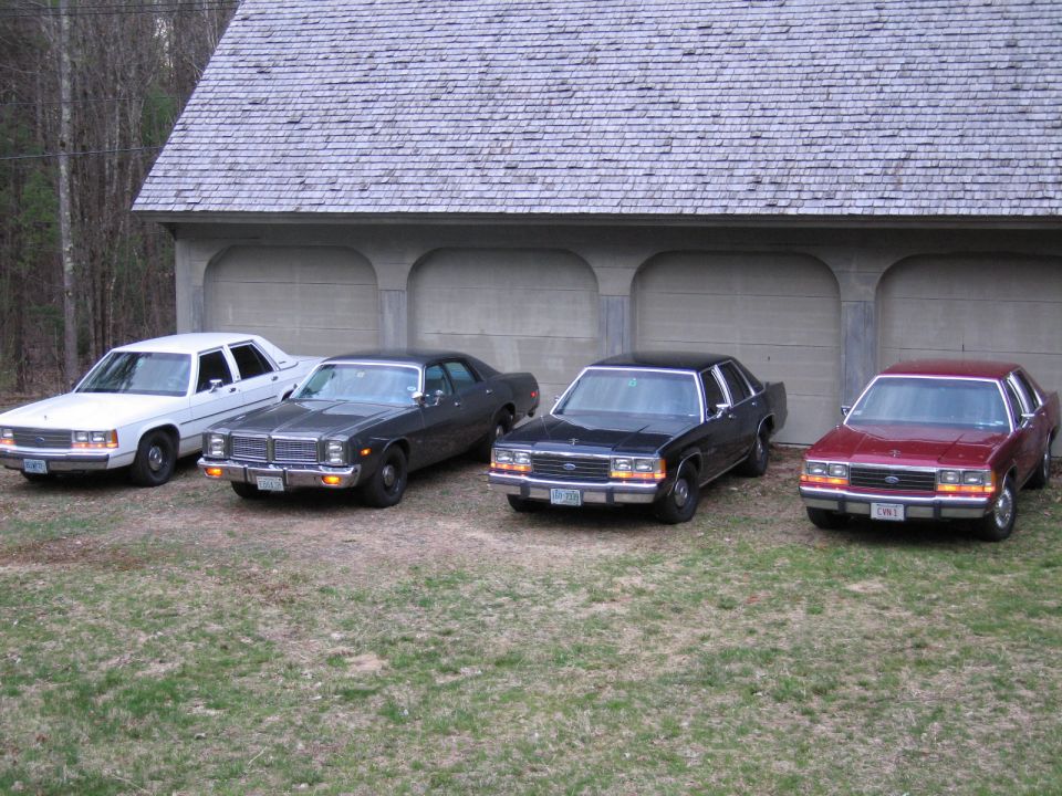 Four sedans parked in front of a garage. The white car on the left appears to be a Ford Crown Victoria. The other three cars are dark colored sedans