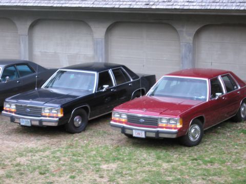 1991 Ford LTD Crown Victoria - Two Ford Crown Victoria LTDs, one red and one black, are parked in front of a garage.