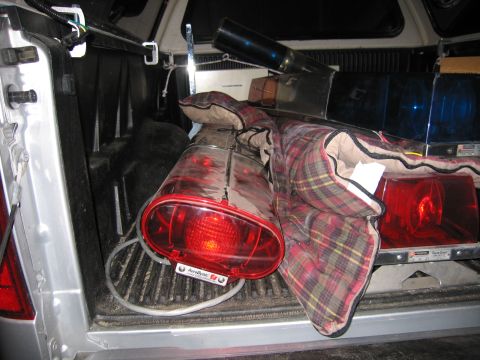1991 Ford LTD Crown Victoria - red emergency beacon light, a chrome siren housing and other gear rest on a plaid blanket inside a pickup truck bed at night.