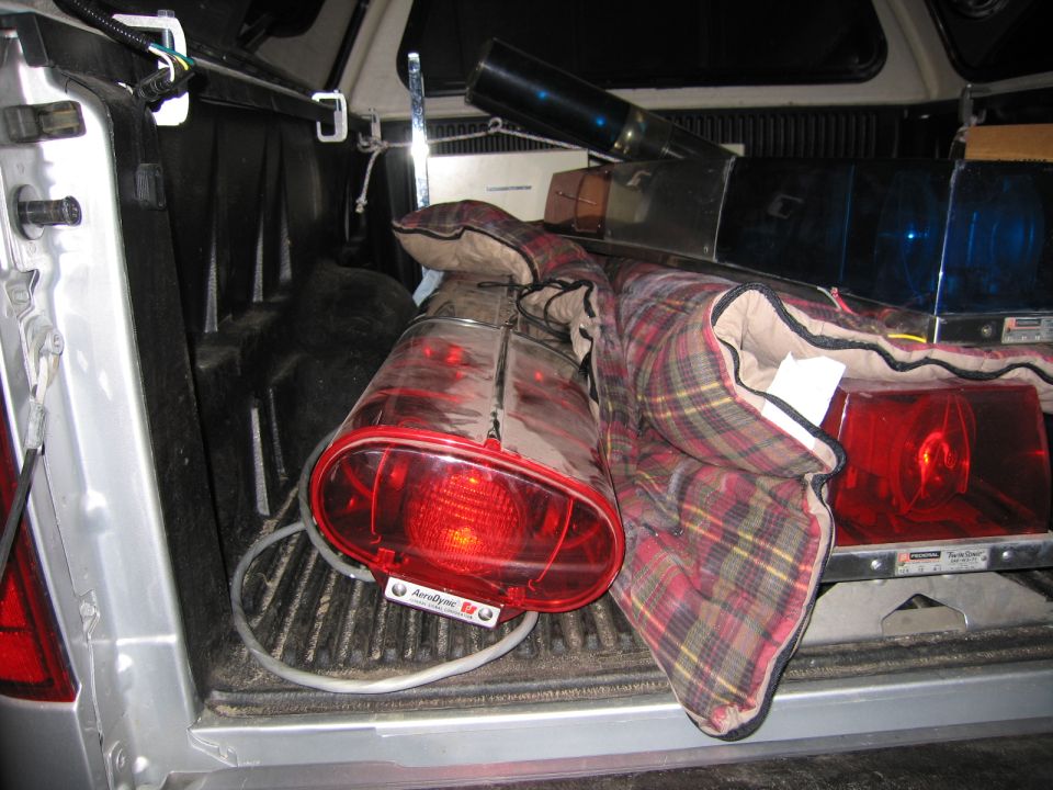 Red emergency beacon light and a chrome siren on a plaid blanket inside a pickup bed