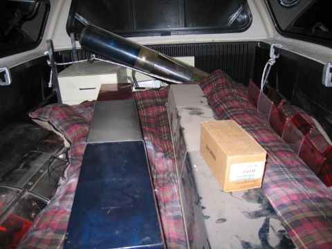 1991 Ford LTD Crown Victoria - Emergency beacon lights, a black spotlight and a cardboard box are bundled in plaid blankets in a pickup truck bed at night.