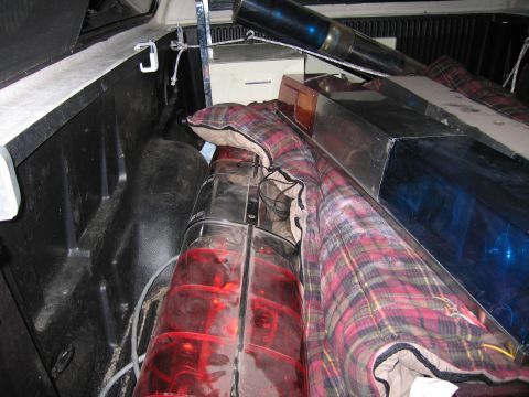 1991 Ford LTD Crown Victoria - Red and blue emergency beacon lights and a black spotlight are wrapped in plaid blankets inside a pickup truck bed at night.