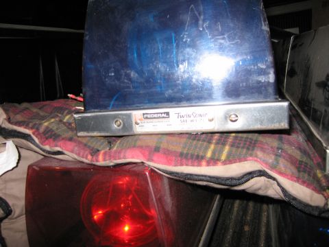 1991 Ford LTD Crown Victoria - Federal Twin Sonic blue emergency beacon sits above a glowing red beacon on a plaid blanket in a dim truck bed.
